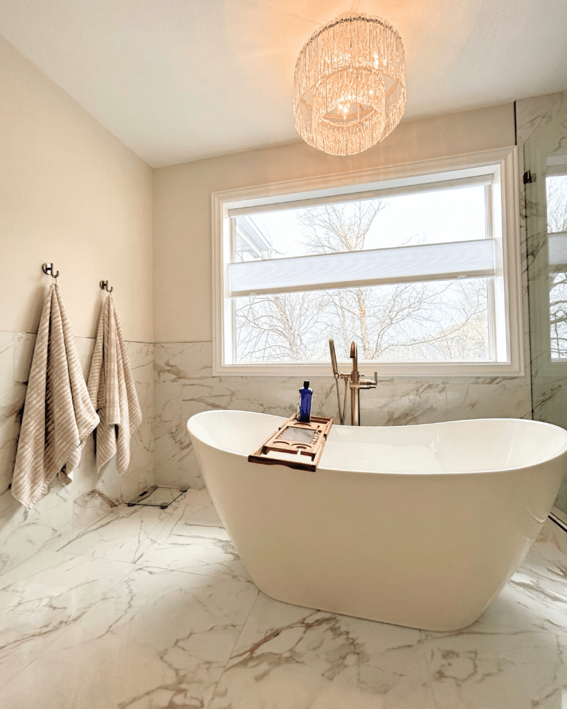Luxurious bathroom featuring a modern white freestanding bathtub on white marble tile flooring, set beneath a sparkling crystal chandelier and large window with soft natural light.