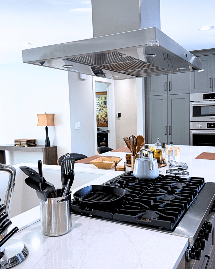 Modern kitchen cooktop on a quartz island beneath a sleek stainless steel range hood, surrounded by light wood and charcoal cabinetry.