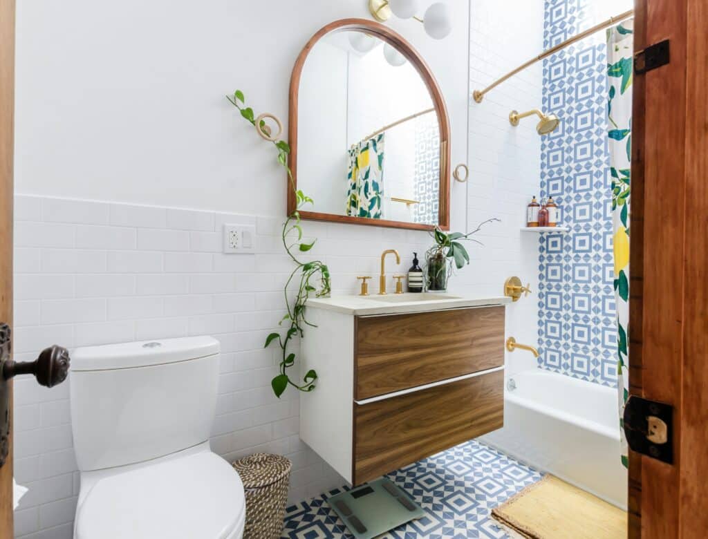 Bright and playful bathroom with blue geometric floor and shower tiles, floating wood vanity, gold fixtures, and leafy green plants adding a touch of nature.
