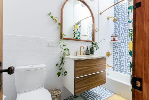 Bright and playful bathroom with blue geometric floor and shower tiles, floating wood vanity, gold fixtures, and leafy green plants adding a touch of nature.